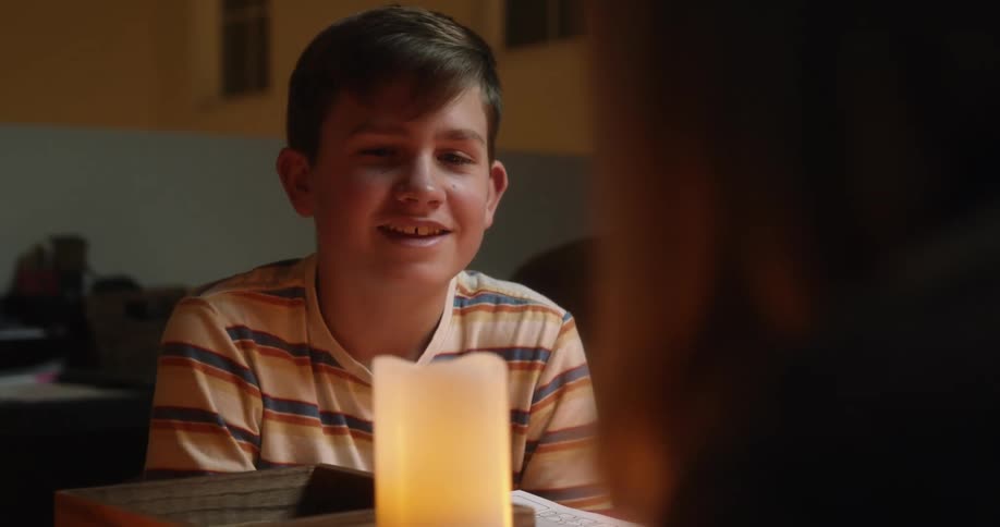 A young boy smiles at his mom who sits at a table lit with candles across from him