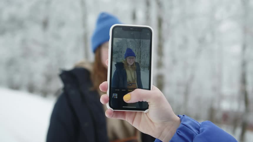 Person uses mobile phone to photograph woman standing in snow covered forest