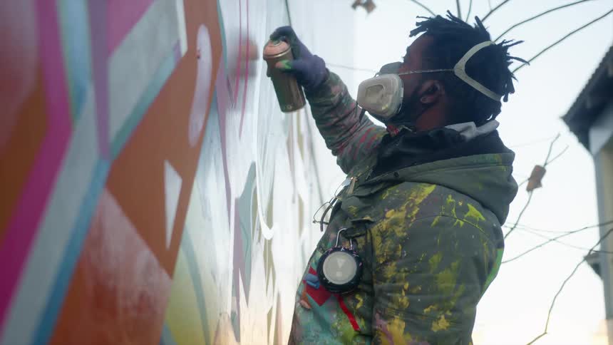 Artist works on mural wall with spray paint near building