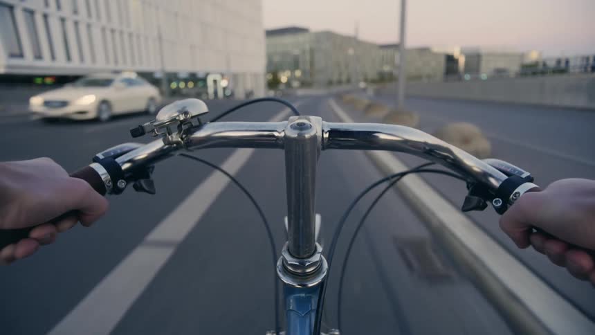 A person riding a bike on a bike lane with buildings the sky and clouds in the background POV