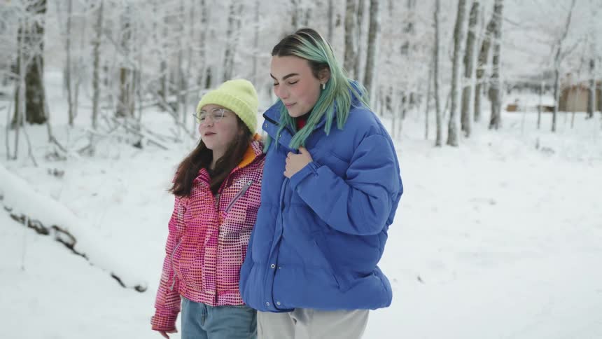 Two girls with coats walking in snow with trees and a house in the background