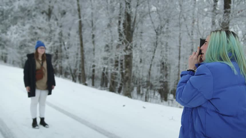A teenage girl stands on a snow covered road next to a forest and poses for a photo as another girl takes it