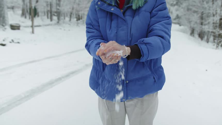 A woman with a coat picking up snow with trees and a road in the background