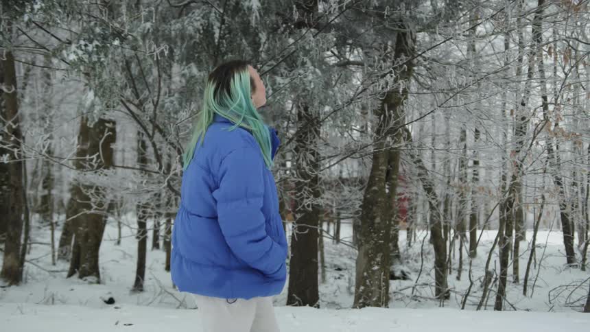 Woman with green hair and wearing a blue jacket walking among snow covered trees looking around