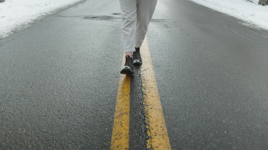 A walk in snow boots and khakis walks down the yellow line of a paved road surrounded by snow