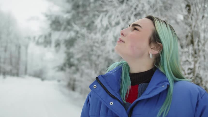 Woman with green dyed hair wearing jacket stands in snow covered forest looking around