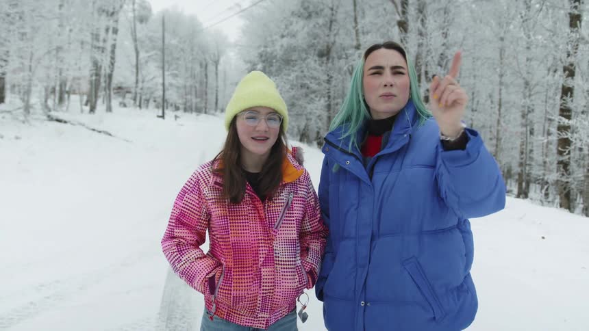Woman points out things to girl as they stand on snow covered road bordered by forest