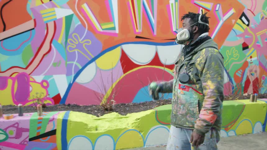 Man wearing protective breathing mask carries spray can past colorful mural wall and flowerbed