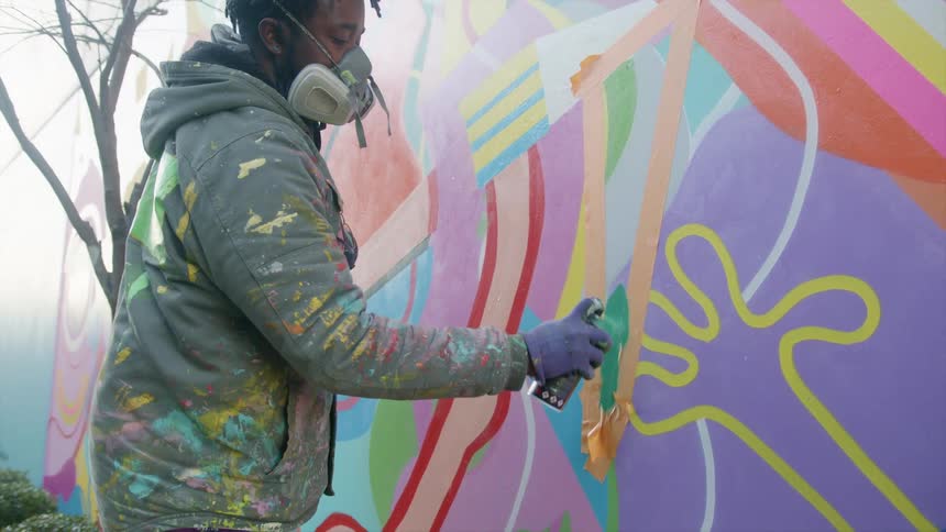A man spray paints a taped off section of a colorful mural