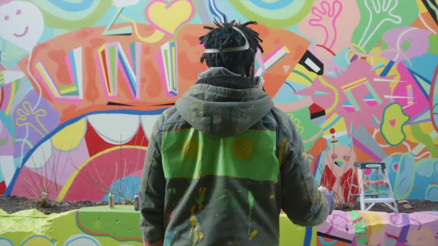 A man with a mask and gloves shaking a spray can with a wall with graffiti in the background