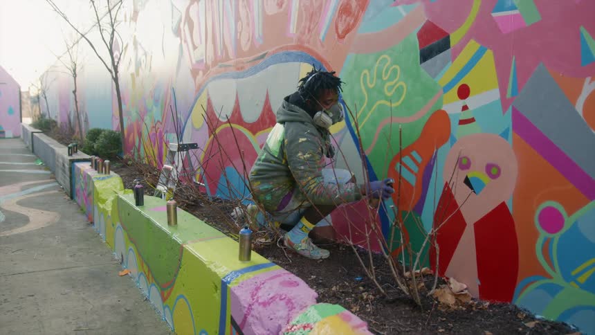 A man in a breathing mask uses spray paint to paint a mural on an outdoor wall during the day