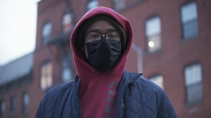 A man poses in front of an apartment building wearing a face mask and hoodie with the hood pulled up