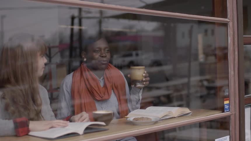 Two women sit in front of the window of a coffee shop with mugs and books