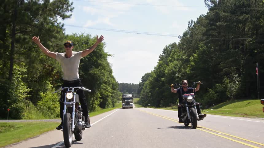 Two men riding motorcycles down a road one stands as he rides