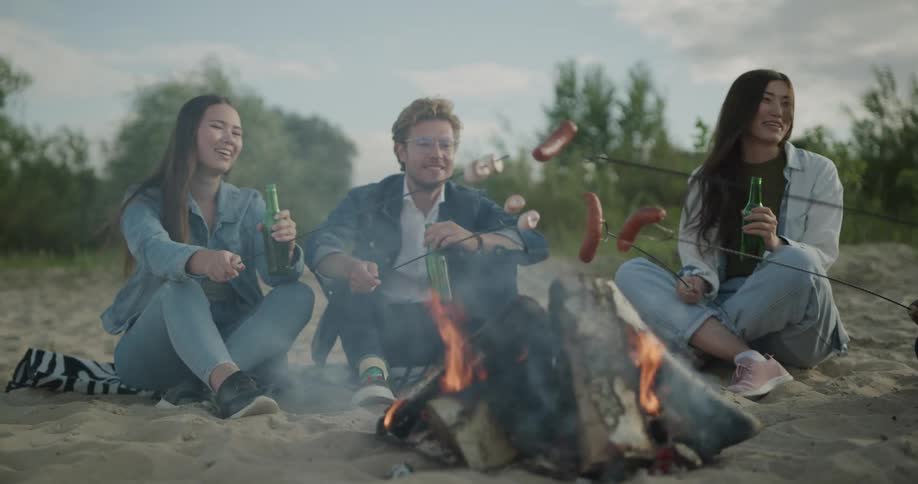 Friends drink beer as they sit around a campfire roasting marshmallows and hot dogs