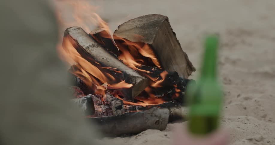 A person holding a beer bottle with sand logs and fire in the background