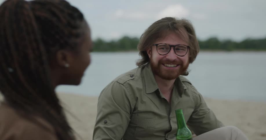 A man and woman clink beer glasses together and drink while sitting on the beach