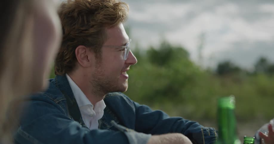 A man sits outside with friend and they talk and drink beer together