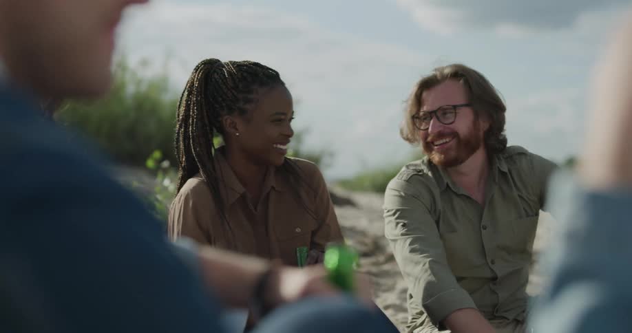 A man and woman sit on the sand with a group of friends drinking talking and laughing