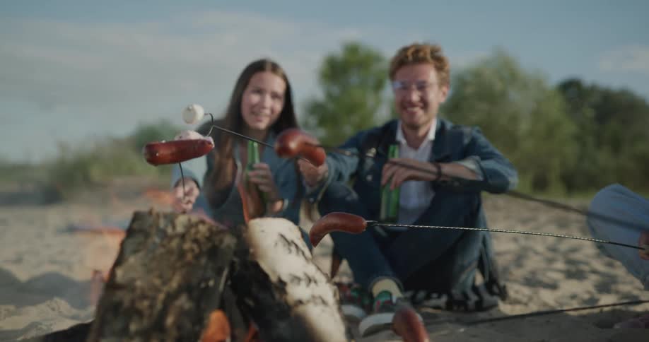 A group of people sit in the sand around a campfire as they roast marshmallows and wieners on skewers