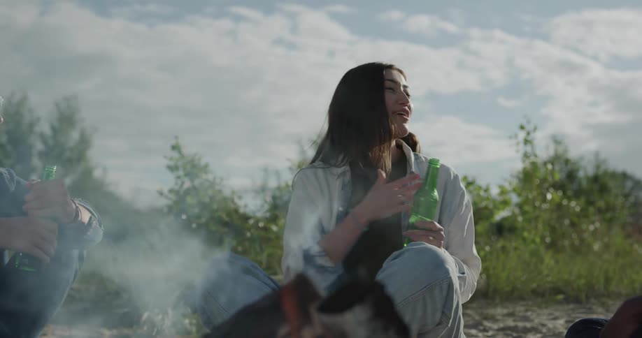 Friends sit around a campfire laughing and drinking beer