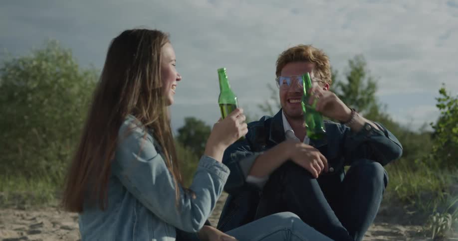 A man and woman clink beers and drink while sitting on the beach around a campfire