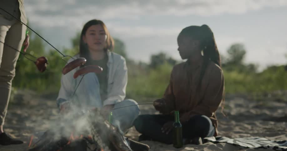 A group of friends talk and laugh as they cook hotdogs over a campfire