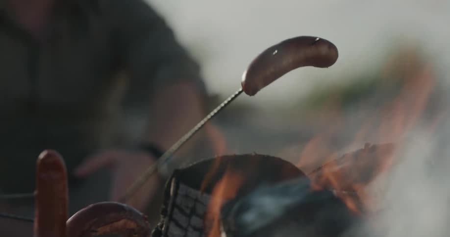 People hold wieners on skewers over a campfire during the day