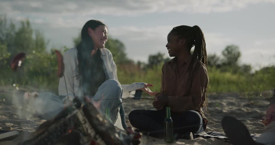 Two female friends sitting on the sand roasting sausages over a campfire laughing and hugging