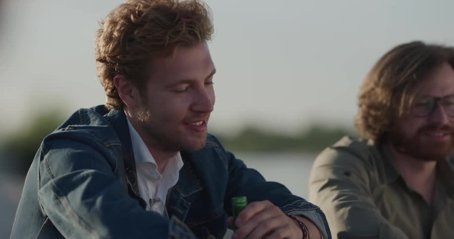 Two men drink beer from green bottles while sitting around a campfire near the ocean