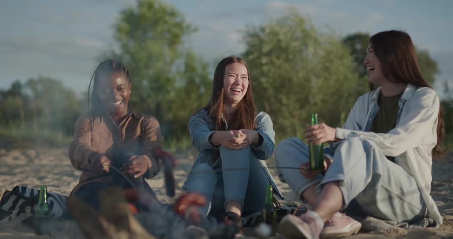 Three women sitting by a campfire roasting sausages with trees the sky and clouds in the background