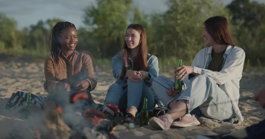 Three women roast wieners over a campfire while they sit on a beach