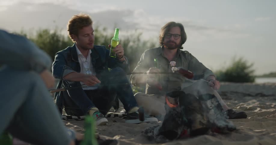 Two men sitting on the shore by a fire and roasting sausages and drinking beer with the sky and clouds in the background