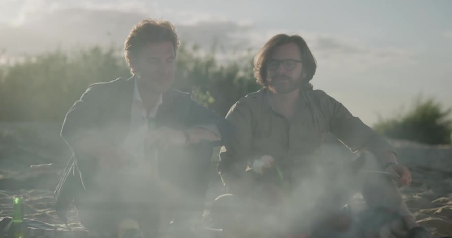 Two men sitting on the shore by a fire drinking beer with the sky and clouds in the background