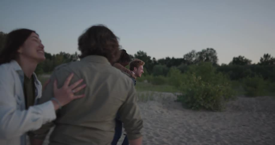 A man gives a woman a piggy back on a beach as they walk with another man and woman that laugh and talk