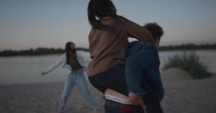A woman on a mans back on a shore with two women and a man the sky and clouds in the background