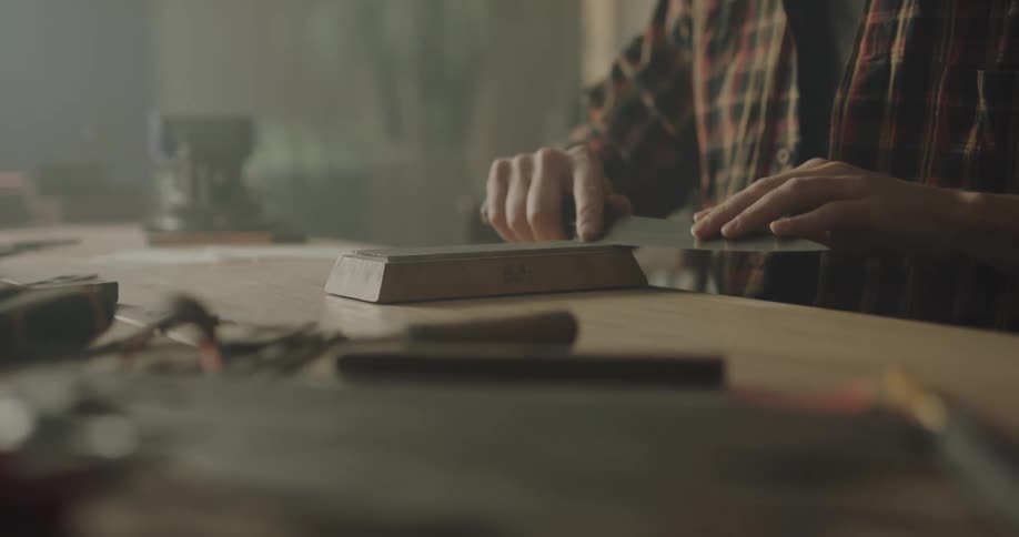 Man sharpening hand made knifes on a wetstone over a workbench in a workhsop