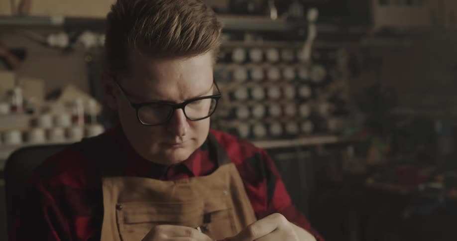 A man in flannel and an apron sits at a workbench sewing leather held in a vise