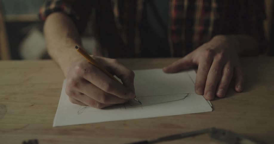A person in a workshop sitting at a desk drawing a knife on a paper with a pencil