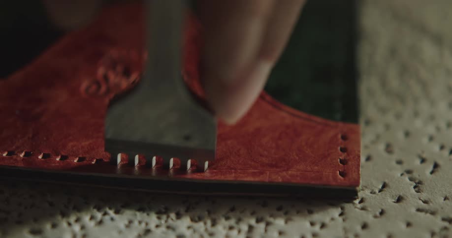 A man uses a fork like tool to put holes along the edges of a piece of leather