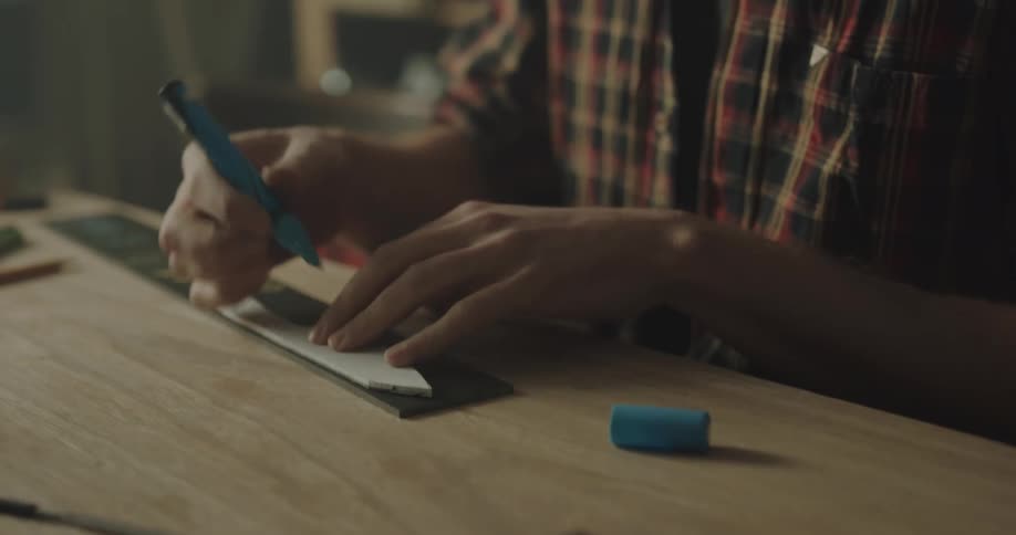 A man in a workshop sitting at a table outlining a piece of wood with a marker