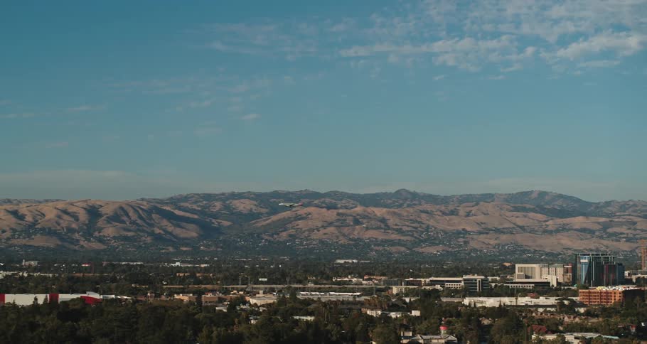 An airplane flying with trees and buildings in the foreground and a mountain range the sky and clouds in the background