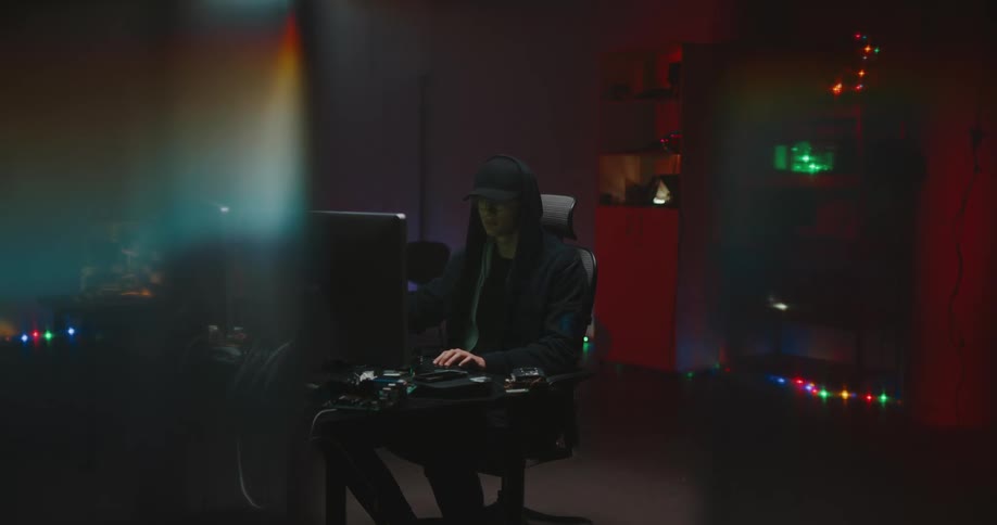 A man wearing his hoodie over his cap sits at a computer snacking while he plays