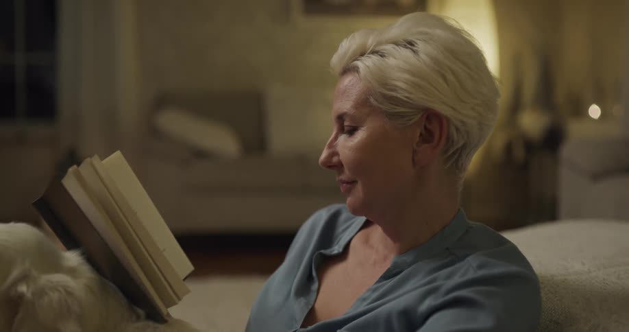 Woman with short grey hair reads book while stroking white dog in the living room at home