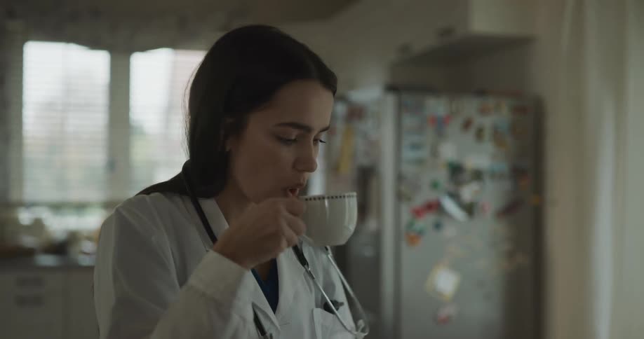 A young woman at home puts a stethoscope around her neck takes a drink of coffee and looks at her cell phone