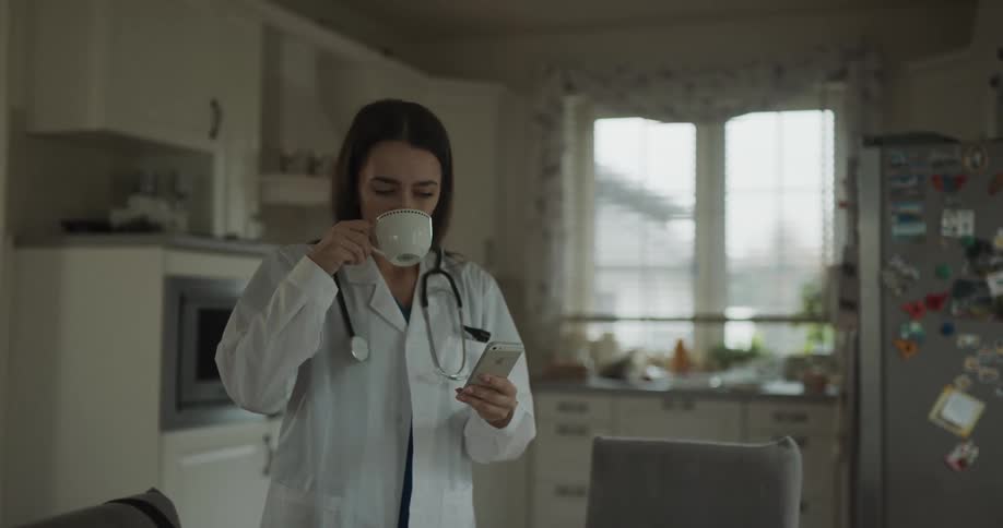 A doctor standing in the kitchen of a house places a stethoscope around her neck before taking a sip of coffee as she checks her phone