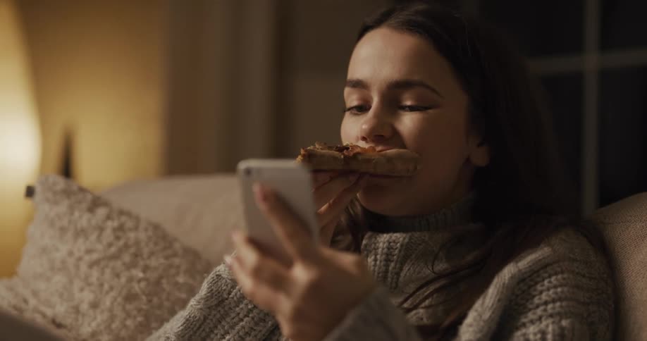 A woman sitting on a couch eating a slice of pizza and texting on her phone
