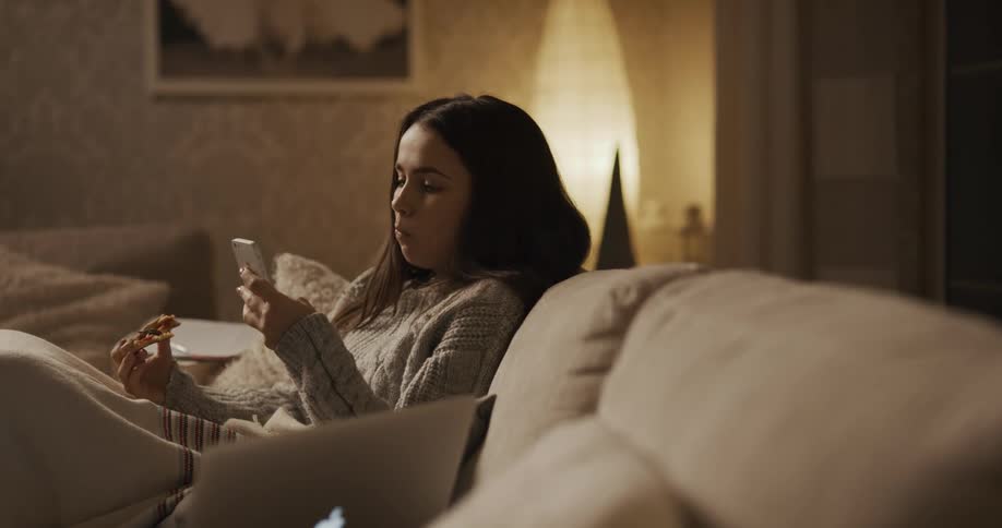 A woman takes a bite of pizza then leans back against the couch cushion texting on her phone