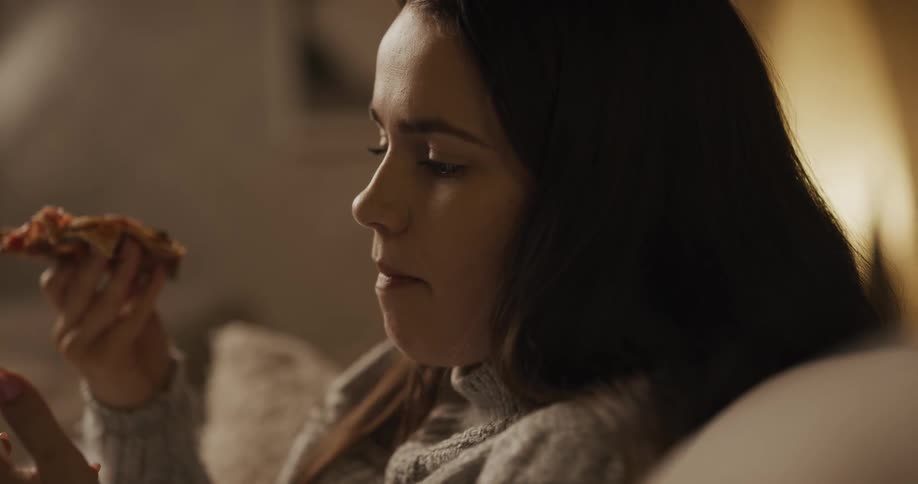 Woman eats a slice of pizza as she sits on a sofa