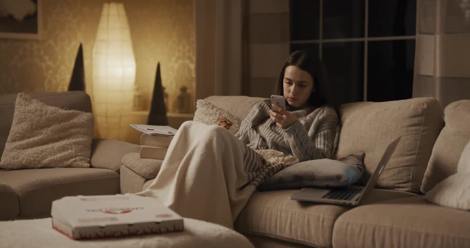 A young woman sitting under a blanket on the sofa in the living room of a house eats pizza while reading her smartphone